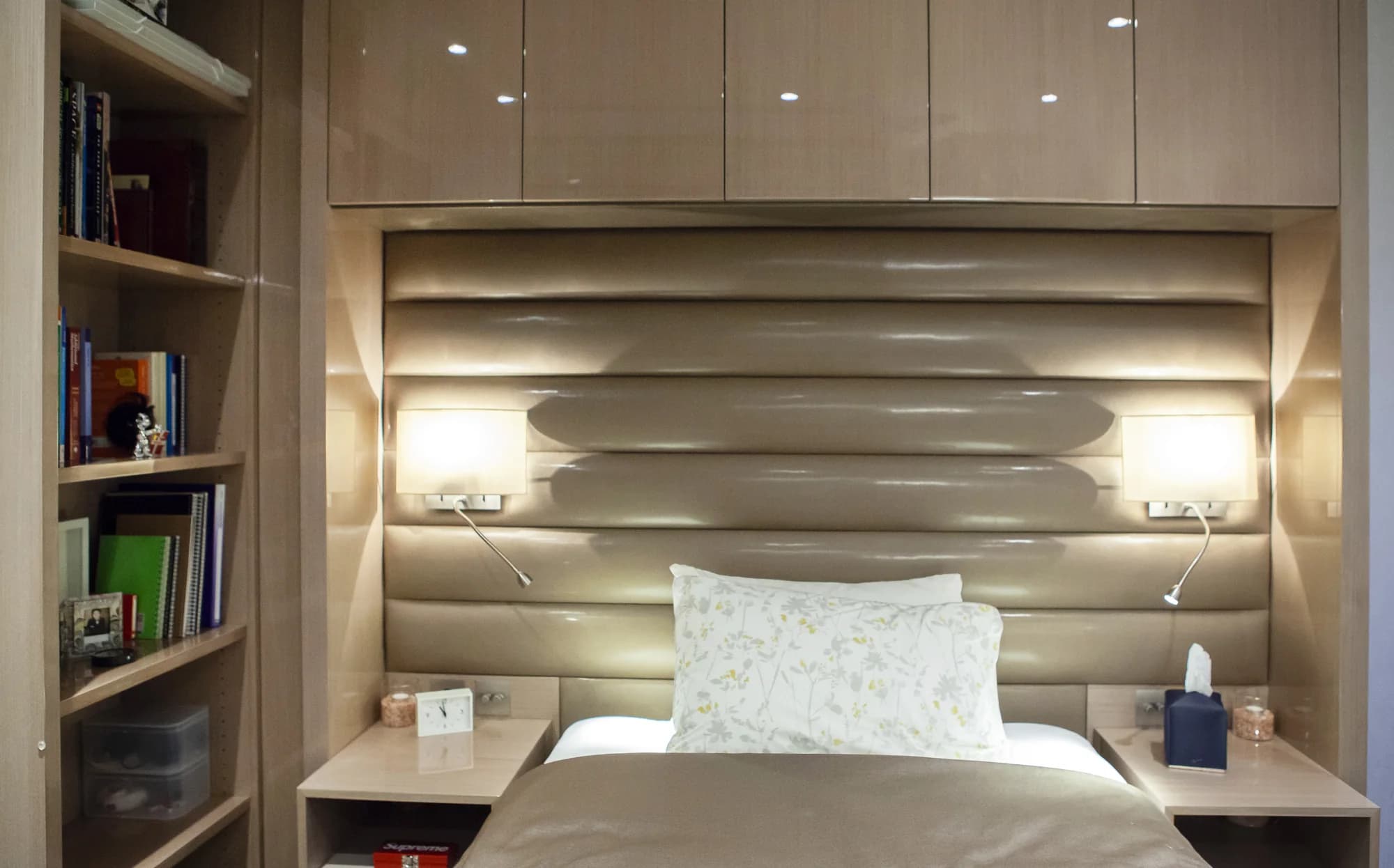 Curved padded headboard beneath glossy overhead cupboards with open book storage alongside
